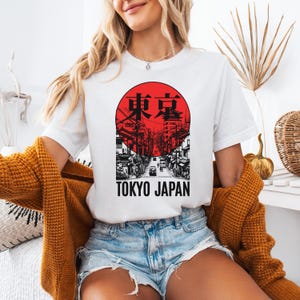 May include: White t-shirt featuring a red and black graphic of a Tokyo street scene within a circle. The text "TOKYO JAPAN" is below the graphic. The shirt is worn with denim shorts and a mustard-colored cardigan.
