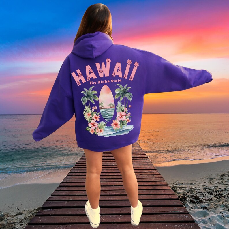 May include: A purple hoodie with a Hawaiian-themed graphic on the back. The graphic includes the word "Hawaii" in pink, palm trees, flowers, and a surfboard design. The text "The Aloha State" is also included.