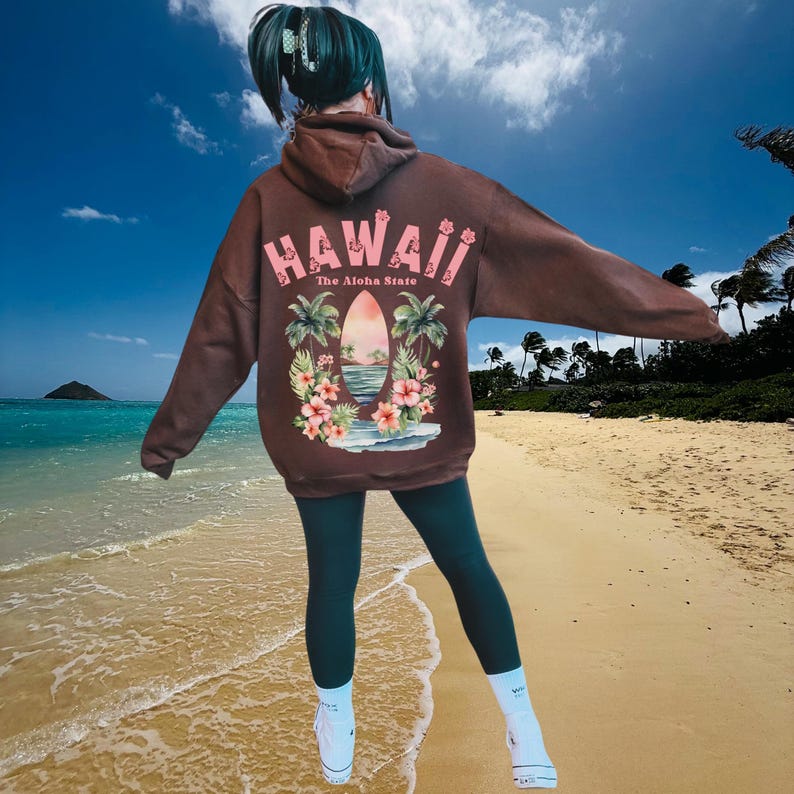 May include: Brown oversized hoodie with the word "HAWAII" in pink, along with a tropical graphic. The graphic includes a surfboard, palm trees, and flowers. The hoodie is worn with dark green leggings and white sneakers.