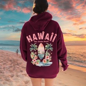 May include: A maroon hoodie with a graphic on the back that says "HAWAII" in pink letters. Below the word "HAWAII" is the phrase "The Aloha State". The graphic also includes a beach scene with palm trees and flowers.