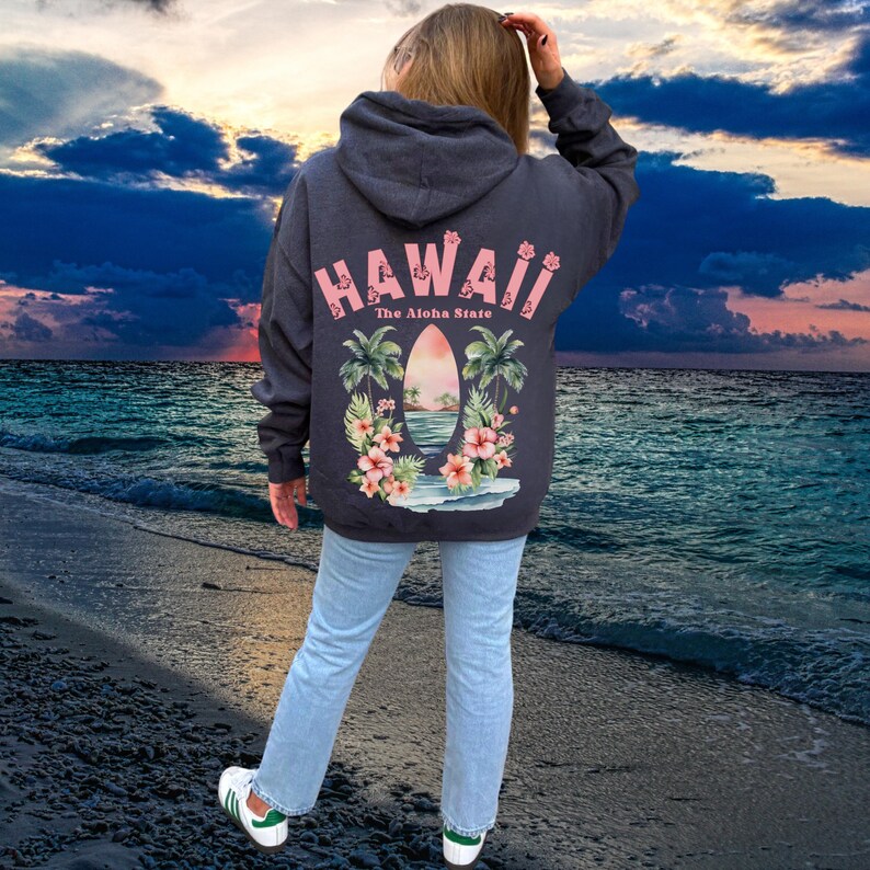 May include: Dark gray hoodie with the word "HAWAII" in pink across the back, along with "The Aloha State." The design includes a surfboard, palm trees, and flowers. The person is wearing light blue jeans and white sneakers.