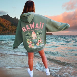 May include: Sage green hoodie with the word "Hawaii" in pink, along with a tropical graphic on the back. The graphic includes palm trees, flowers, and a surfboard. The text "The Aloha State" is also included.