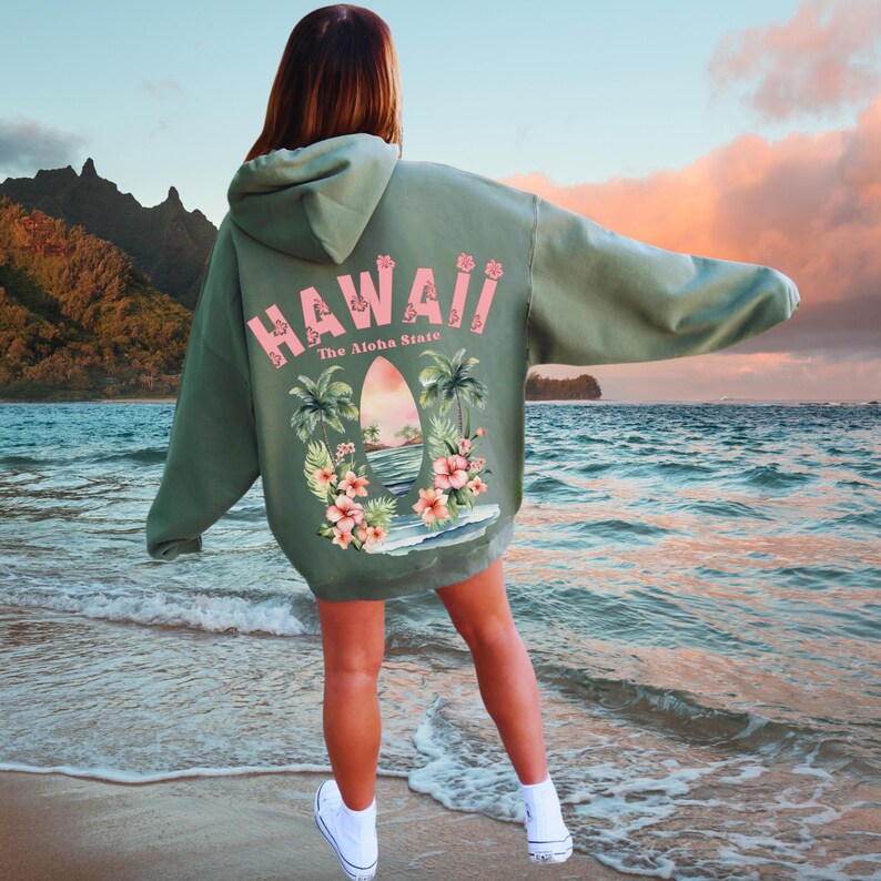 May include: Sage green hoodie with the word "Hawaii" in pink, along with a tropical graphic on the back. The graphic includes palm trees, flowers, and a surfboard. The text "The Aloha State" is also included.