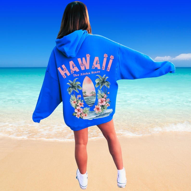 May include: A blue hoodie with a Hawaiian theme. The back of the hoodie features the word "HAWAII" in pink, along with a surfboard, palm trees, and tropical flowers. The text "The Aloha State" is also visible.