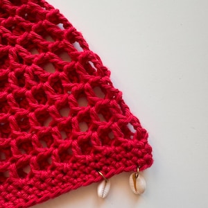 May include: A section of a bright red, open-weave crochet garment. The crochet has a net-like pattern. Two small, white seashells are attached to the bottom edge with gold-colored rings. The background is plain white.