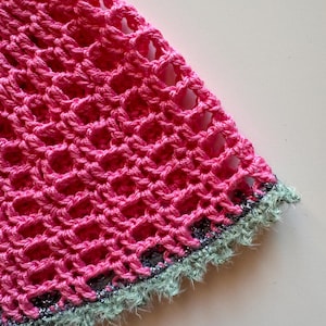 May include: Close-up of a section of a crocheted item. The main color is a vibrant pink, with a mesh-like pattern. The edge is trimmed with a light green, fuzzy border, and a thin silver thread. The texture is visible.
