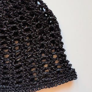 May include: Close-up of a black crocheted hat with a textured, open weave design. The hat is made of a shiny, dark yarn, and the edges are slightly scalloped. The background is a plain white surface.
