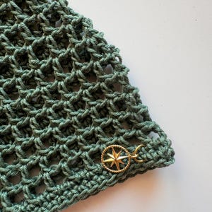 May include: Close-up of a section of a green crocheted item. The open weave pattern is visible. A gold-colored charm in the shape of a compass rose is attached to the edge of the item.