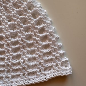 May include: Close-up of a white, open-weave crochet fabric. The texture is visible, with a repeating pattern of interconnected loops. The edge is finished with a solid, tightly crocheted border. The fabric is likely used for clothing or accessories.
