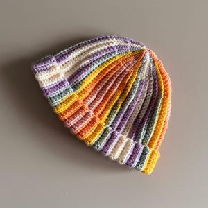 May include: A handmade, colorful crochet beanie hat. The hat features vertical stripes in a rainbow of colors, including yellow, orange, purple, and white. The brim is folded up, and the hat is made of a soft, textured yarn.