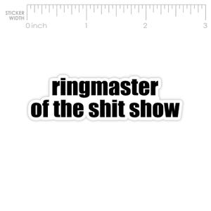 May include: A white sticker with the text "ringmaster of the shit show" in black, bold font. The sticker has a black border. A ruler in inches is visible at the top, with the equivalent in cm.