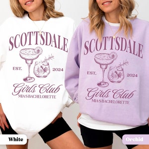 Puede incluir: Dos sudaderas, una blanca y otra morada, con el texto "Scottsdale Est. 2024 Girls Club Mia's Bachelorette" y un gr&aacute;fico de dos c&oacute;cteles. La sudadera de la izquierda es blanca y la sudadera de la derecha es morada.