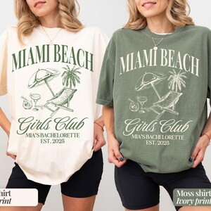 May include: Two t-shirts with a beach scene and the text "Miami Beach Girls Club Mia's Bachelorette Est. 2025". One shirt is ivory with moss green print and the other is moss green with ivory print.