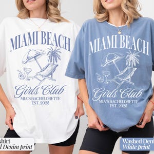May include: Two t-shirts with a graphic print of a beach scene with a lounge chair, palm trees, and the text "Miami Beach Girls Club Mia's Bachelorette Est. 2025". One shirt is white with a blue print and the other is a washed denim blue with a white print.