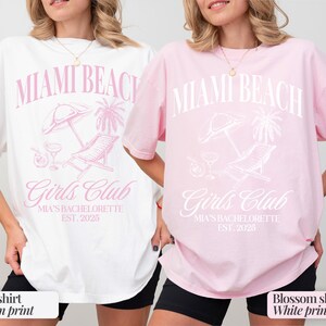 May include: Two white t-shirts with a pink graphic print that says "Miami Beach Girls Club Mia's Bachelorette Est. 2025". One shirt is white with pink print and the other is pink with white print.