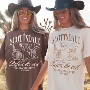 May include: Two white t-shirts with a brown graphic design. The graphic features a cocktail glass, a cowboy hat, and cowboy boots. The text on the shirts reads "Scottsdale Before the Veil" and "Mia's Bachelorette Est. 2024".