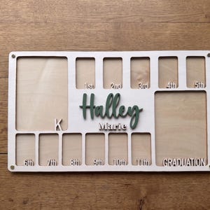May include: A rectangular light wood photo collage frame featuring multiple cutouts for school years labeled 1st through 11th, plus Graduation. The center displays the name Halley Marie in raised dark green and white lettering with a letter K.