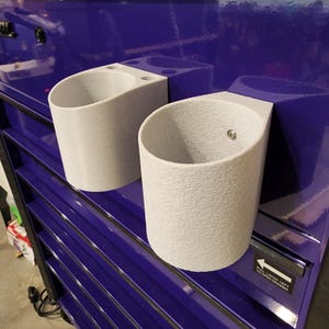 May include: Two grey plastic cup holders mounted on a purple metal toolbox. The cup holders have a textured surface and are designed to hold cups or other small items.