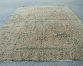 May include: A large rectangular area rug featuring a faded beige background with intricate floral and vine patterns in muted green and brown tones. The design includes a central medallion motif surrounded by a detailed border with repeating floral elements.