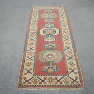 Könnte beinhalten: Ein rechteckiger Läuferteppich mit einem roten Mittelfeld mit geometrischen Designs in Beige, Blau und Grün. Der Rand ist beige mit sich wiederholenden Blumen- und Kreuzmustern. Der Teppich liegt auf einer grauen Oberfläche.