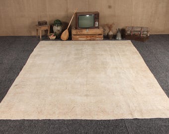 May include: A large, rectangular area rug featuring a neutral, off-white and beige color palette with a subtle, faded pattern across its surface. The rug is laid flat on a dark gray floor, showcasing a low-pile texture and a worn, vintage appearance.