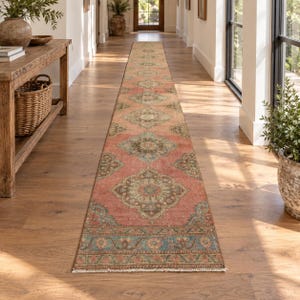 May include: A long, narrow runner rug featuring a repeating medallion pattern in shades of coral, sage green, and muted blue. The rug has a distressed finish with a decorative border and short, light-colored fringe at the ends, laid flat on a floor.