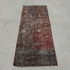 Puede incluir: Una alfombra rectangular roja desgastada con un intrincado diseño floral en blanco y gris. La alfombra tiene una apariencia descolorida, con tonos más oscuros de rojo y marrón en los bordes.