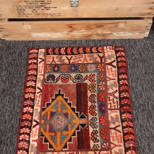 Könnte beinhalten: Ein kleiner, rechteckiger Teppich mit einem geometrischen Muster in Rot-, Braun-, Beige- und Blautönen. Der Teppich hat ein zentrales Rautenmuster und florale Motive. Der Teppich liegt auf einer grauen Oberfläche.