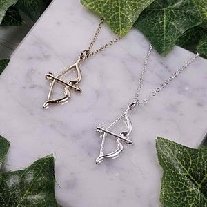 May include: Two necklaces with bow and arrow pendants. One necklace is gold and the other is silver. The necklaces are displayed on a white surface with green leaves.