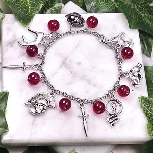 Puede incluir: Un brazalete de plata con cuentas de vidrio rojas y varios dijes de plata, incluyendo una espada, una serpiente, una cabeza de ciervo, un casco y una cabeza de lobo.