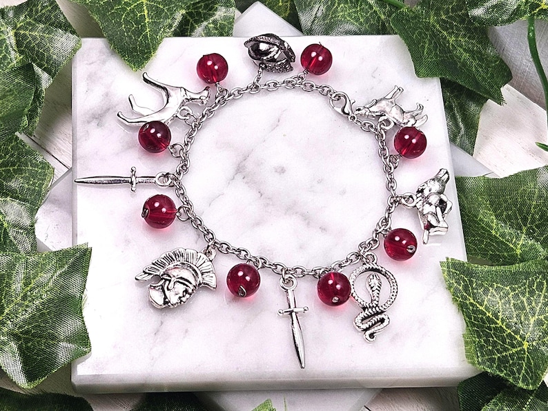 Puede incluir: Un brazalete de plata con cuentas de vidrio rojas y varios dijes de plata, incluyendo una espada, una serpiente, una cabeza de ciervo, un casco y una cabeza de lobo.