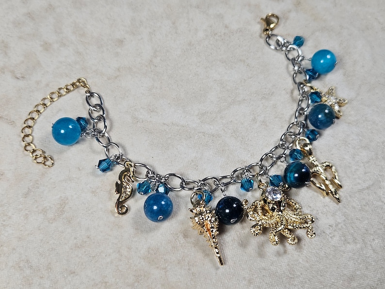 May include: Silver-tone charm bracelet featuring blue beads and gold-tone charms. Charms include a seahorse, starfish, and other sea-themed designs. The bracelet has an adjustable chain for a comfortable fit.