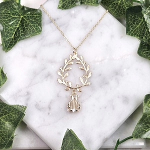 Puede incluir: Collar de oro con un colgante de corona y un dije de lira. La corona está hecha de hojas delicadas y la lira está detallada con cuerdas.