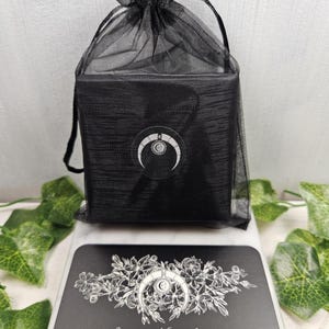 Puede incluir: Una caja negra con un dise&ntilde;o de luna creciente est&aacute; envuelta en una bolsa de malla negra. La caja est&aacute; sobre una tarjeta negra con un dise&ntilde;o floral y el texto "Jennifer Nicole Jewelry".