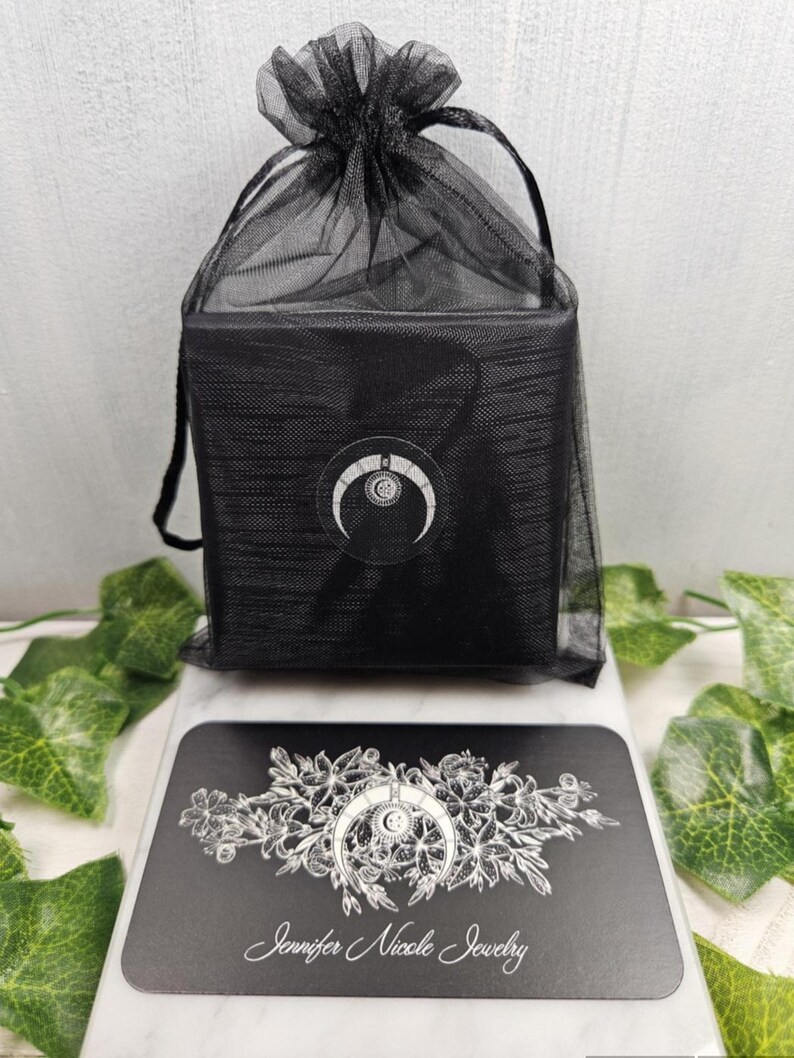 Puede incluir: Una caja negra con un dise&ntilde;o de luna creciente est&aacute; envuelta en una bolsa de malla negra. La caja est&aacute; sobre una tarjeta negra con un dise&ntilde;o floral y el texto "Jennifer Nicole Jewelry".