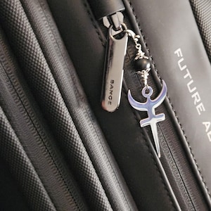 May include: A black zipper pull with a silver charm attached. The charm features a crescent moon and a stylized symbol with a sword. The zipper pull has the word "BANGE" engraved on it.