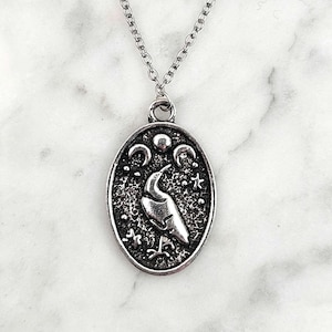 Puede incluir: Collar con colgante plateado. El colgante ovalado presenta una silueta de pájaro, lunas crecientes y estrellas sobre un fondo texturizado. La cadena y el cierre son delicados.