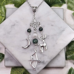 May include: A silver-toned necklace with a decorative pendant featuring a crescent moon, deer, and bow and arrow charms. The necklace also includes green and white beads, and a floral design at the top.
