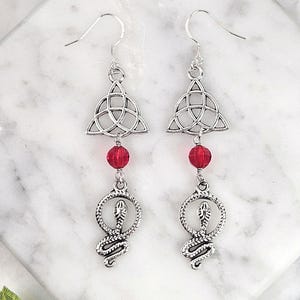 May include: Silver earrings with a red crystal bead and a snake charm hanging from a Celtic knot design.