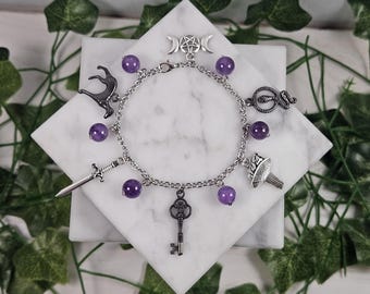 May include: A silver charm bracelet featuring amethyst beads and various charms, including a key, sword, snake, and mushroom. The bracelet also has a triple moon pendant and a black silhouette of a deer.
