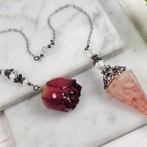 Op de afbeelding: Een zilveren ketting met een roze kristallen slinger en een rode rozenknop hanger. De slinger is een puntig kristal met een zilveren metalen top. De rozenknop is donkerrood en heeft een zilveren metalen lus.