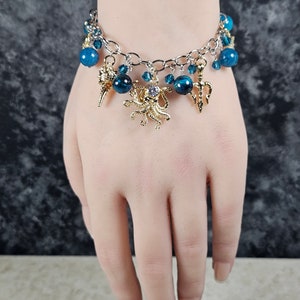 May include: Silver-toned bracelet with blue beads and gold charms. The charms include a seashell, a trident, and an octopus with a central jewel. The bracelet is displayed on a person's wrist.
