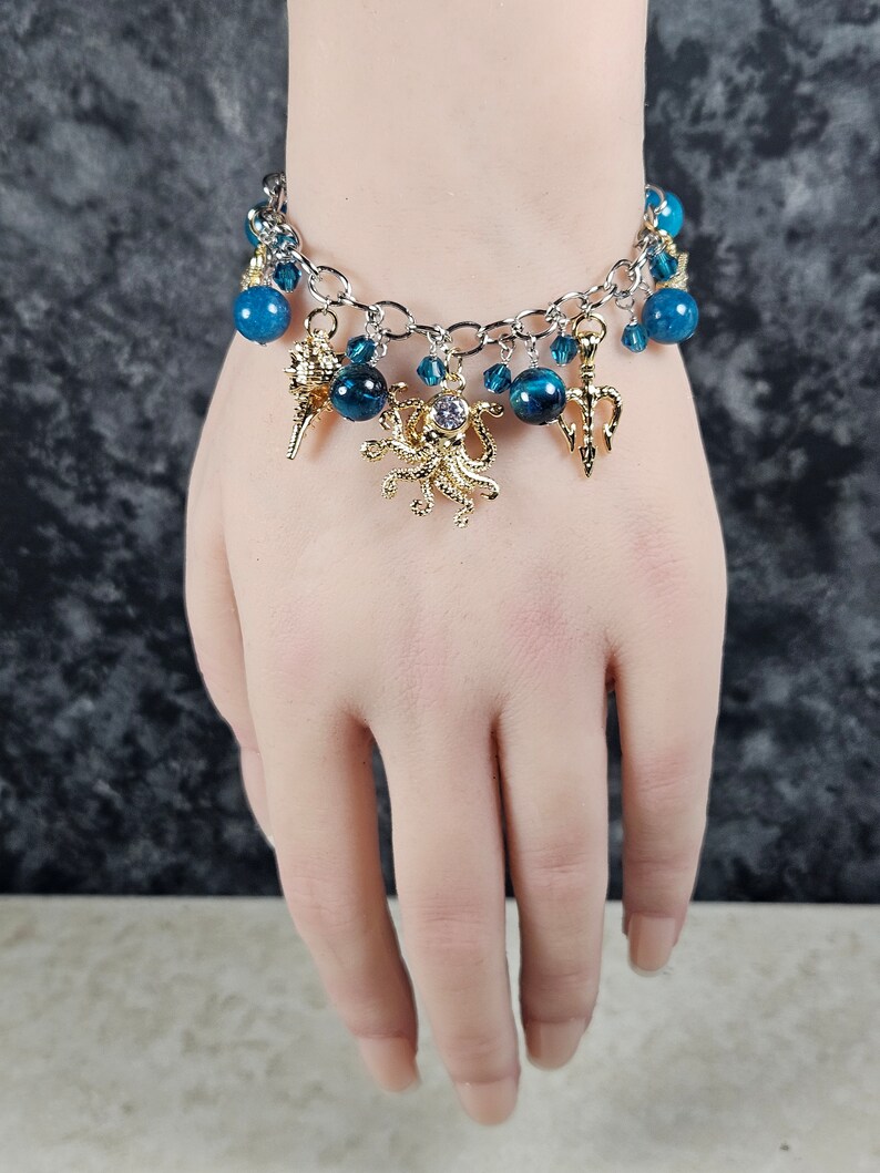 May include: Silver-toned bracelet with blue beads and gold charms. The charms include a seashell, a trident, and an octopus with a central jewel. The bracelet is displayed on a person's wrist.