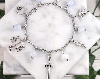 May include: A silver-toned charm bracelet featuring a chain design with various charms. The charms include a sword, wings, a feather, a leaf, and a scale. The bracelet also has light blue and white round beads.