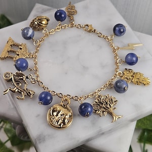 May include: Gold-toned charm bracelet featuring blue stone beads and various charms. Charms include a snake, owl, tree, lightning bolt, and a coin with an image of two animals. The bracelet is displayed on a white marble surface.