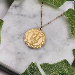 May include: Gold necklace with a round pendant featuring a trident design. The pendant has a decorative border of waves.