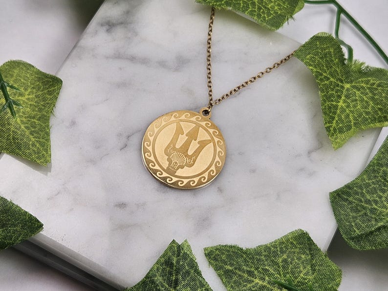 May include: Gold necklace with a round pendant featuring a trident design. The pendant has a decorative border of waves.
