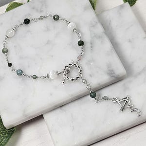 May include: A silver chain bracelet with a silver toggle clasp and a silver charm in the shape of an arrow. The bracelet is adorned with white and green gemstone beads.