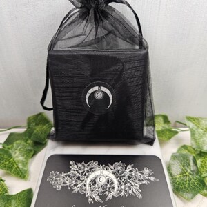 May include: A black gift bag with a drawstring closure, containing a black box. The box has a white crescent moon logo. A black business card with the text "Jennifer Nicole Jewelry" and a floral design is in front of the bag.