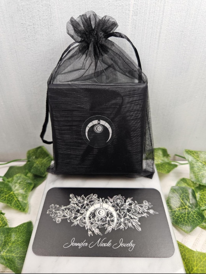 May include: A black gift bag with a drawstring closure, containing a black box. The box has a white crescent moon logo. A black business card with the text "Jennifer Nicole Jewelry" and a floral design is in front of the bag.